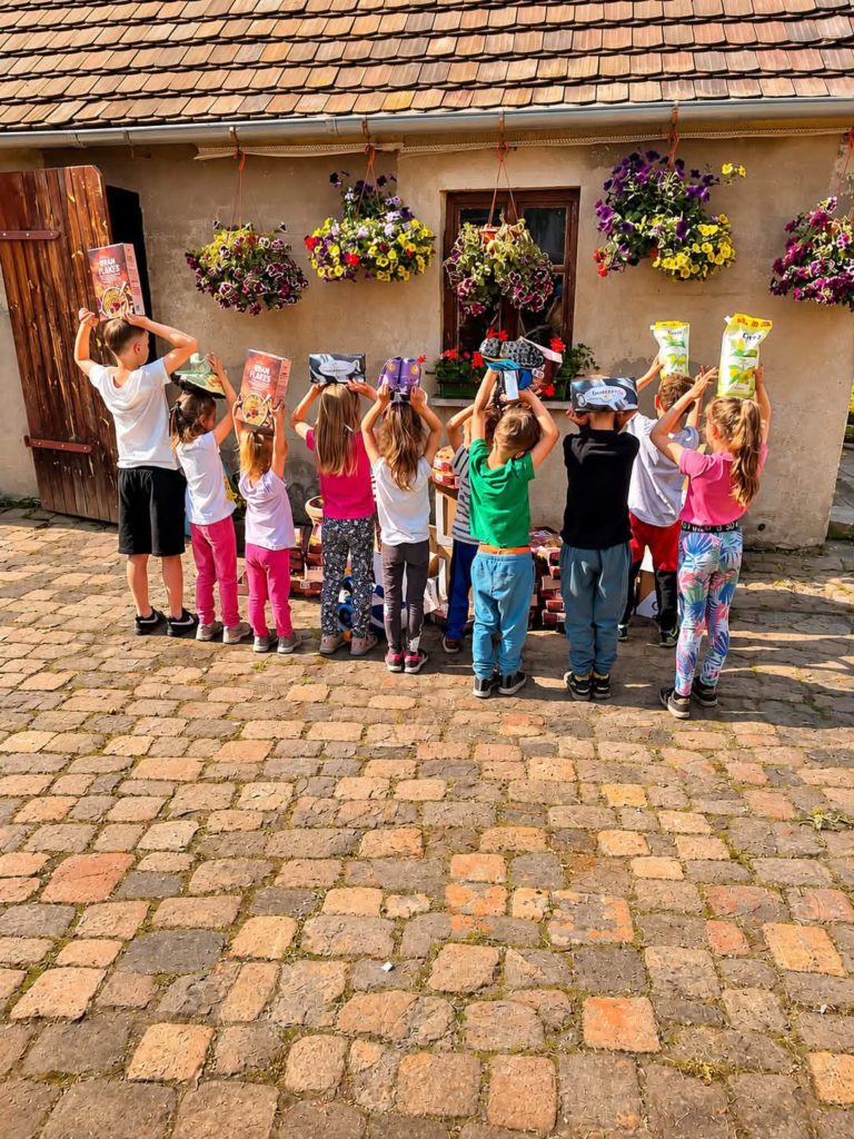 Grupa dzieci stoi przed domem tyłem do obiektywu trzymając w górze w rękach paczki.