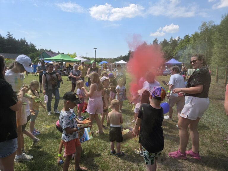 Zdjęcie przedstawia plenerowy festyn rodzinny w słoneczny dzień, podczas którego dzieci i dorośli bawią się kolorowymi proszkami Holi. Na trawiastym placu, w otoczeniu drzew i namiotów, wśród zgromadzonych unosi się duża chmura czerwono-różowego pyłu a uczestnicy mają ślady kolorów na ubraniach i ciele. Na pierwszym planie widać dzieci oraz uśmiechniętą kobietę w okularach przeciwsłonecznych, która obserwuje zabawę.